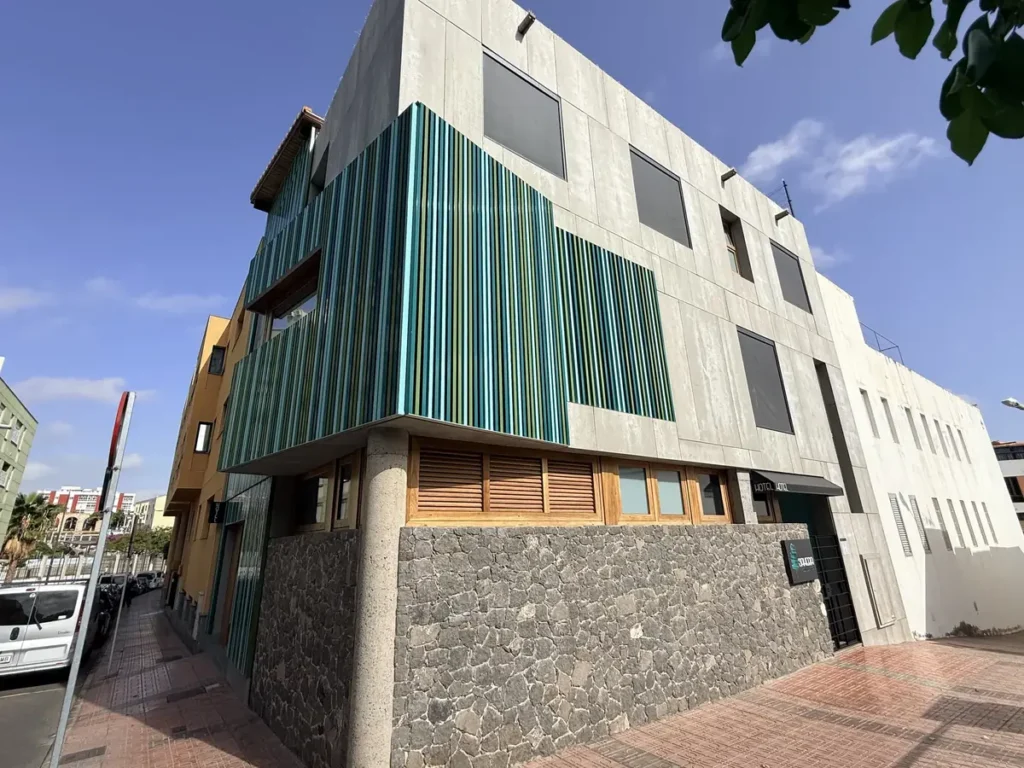 Fachada de SAHERA en esquina, con lamas verdes, madera y piedra volcánica bajo el cielo azul