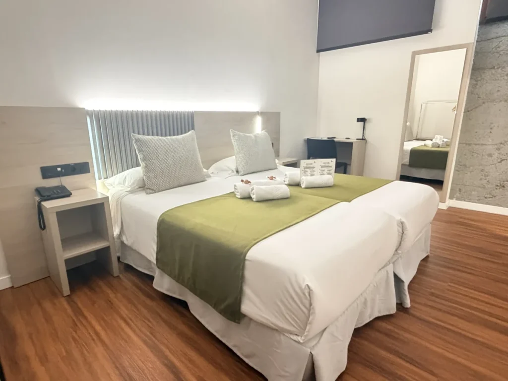 Habitación 101 adaptada: dos camas individuales unidas con colcha verde y toallas, cabecero con luz