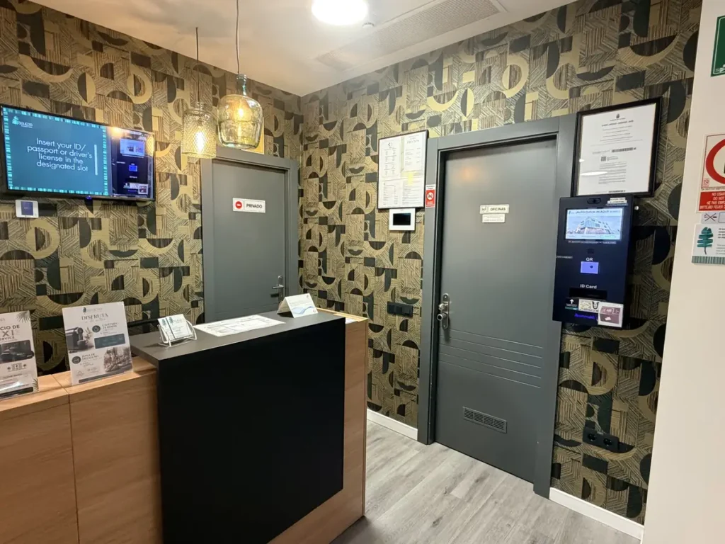 Mostrador de recepción de SAHERA con papel pintado geométrico y, junto a la puerta, el tótem de auto check-in