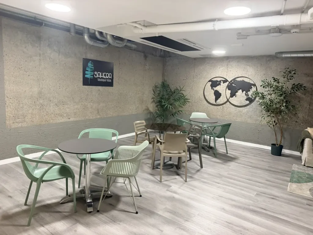 Mesas con sillas verdes y de madera en el salón de usos múltiples, con plantas y el logotipo del hotel